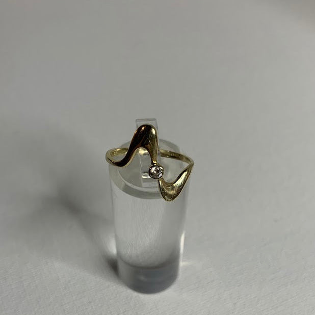 A gold ring with a wavy, organic shape and a small clear gemstone, displayed on a clear cylindrical stand against a white textured background.