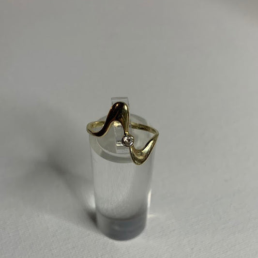 A gold ring with a wavy, organic shape and a small clear gemstone, displayed on a clear cylindrical stand against a white textured background.