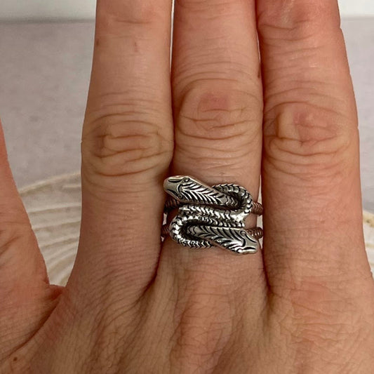 Hand wearing a silver snake with two heads feather-patterned ring on a neutral background